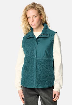 Frau mit lockigem blondem Haar, die eine türkisfarbene ärmellose Fleeceweste über einem cremefarbenen Langarmshirt und dunklen Hosen trägt, steht vor weißem Hintergrund.