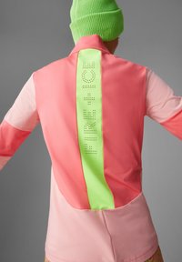 Rosa och grön atletisk jacka med hög krage, försedd med en reflekterande grön baksida som visar "FIRE + ICE" i perforerat skrivtecken.