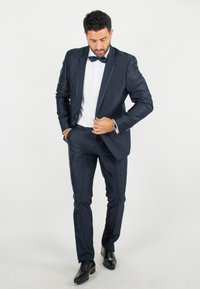 Costume ajusté bleu marine avec un revers châle et une chemise blanche. Accompagné d'un nœud papillon noir et de chaussures de ville noires.