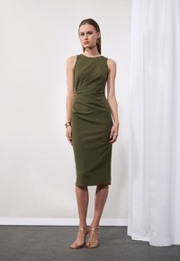 Rochie midi fără mâneci, de culoare verde măsliniu, cu o siluetă ajustată și drapaje subtile, asortată cu sandale aurii cu bretele.