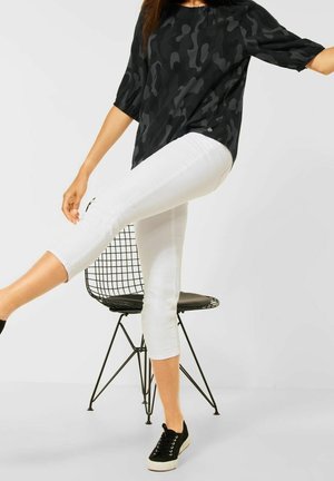 Haut noir à motifs avec des manches trois-quarts associé à un pantalon blanc coupé. Baskets noires. Le modèle pose près d'une chaise en fil moderne.
