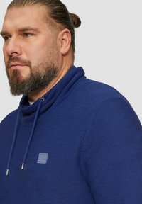 Pulower w kolorze niebieskim kobaltowym z kołnierzem w formie lejka, teksturowanym splotem i naszywką z logo. Posiada sznurki z metalowymi końcówkami, co podkreśla jego design.