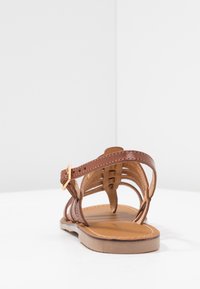 Infradito in pelle marrone con design a strisce incrociate, cinturino alla caviglia regolabile con dettagli dorati e suola piatta color tan.