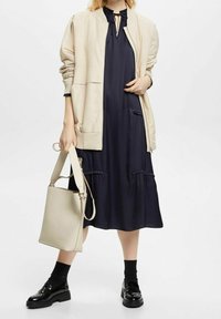 Beige bomberjacka över en marinblå klänning med volangdetaljer, i kombination med en ljus tan-handväska och svarta platåskor med en glänsande finish.