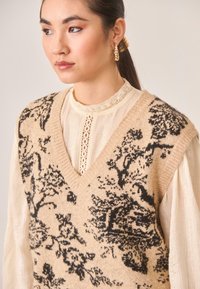 Chaleco de punto beige con patrones florales negros, diseño de escote en V, llevado sobre una blusa de textura crema con cuello de encaje y detalles de botones.