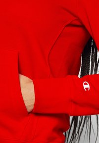 Czerwona bluza z ribbowymi ściągaczami i kieszenią typu kangur. Posiada biały logo na ściągaczu oraz teksturowany materiał. Zbliżenie.