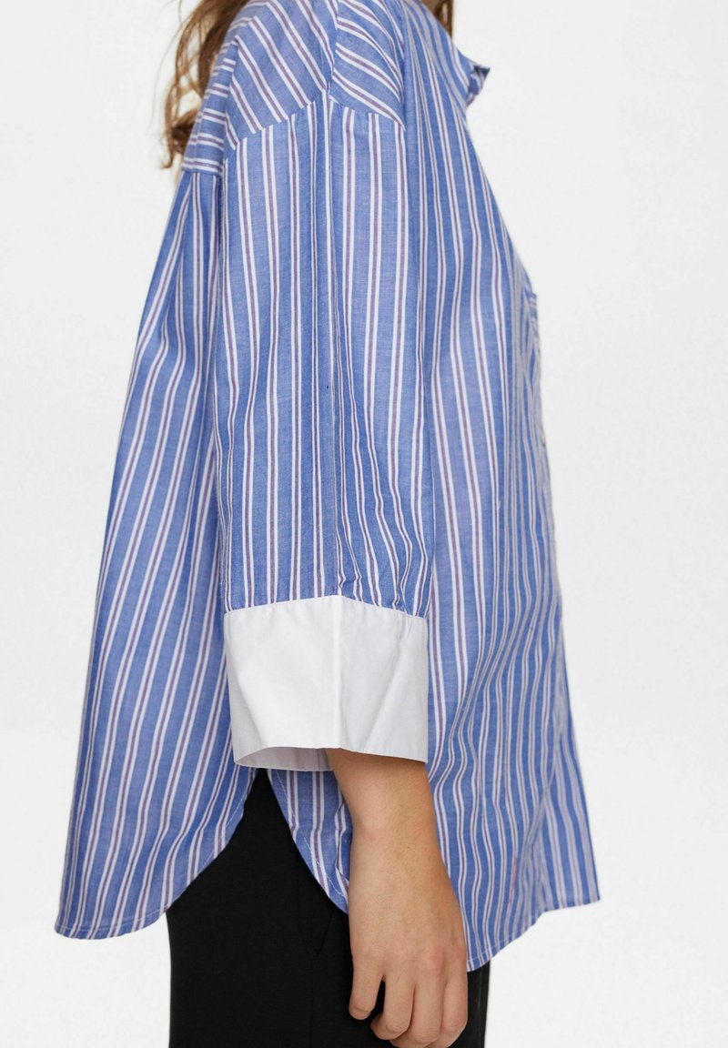 Camicia a righe blu e bianche con colletto abbottonato, maniche lunghe, polsini bianchi e un leggero fondo scampanato. Realizzata in tessuto leggero.