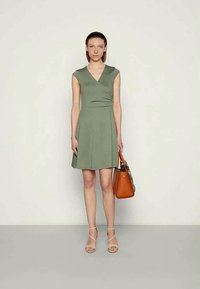 Vestido verde envelopado com mangas curtas, corpete ajustado e saia rodada. Acompanhado por uma mala castanha e sandálias nudas com tiras.