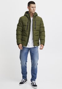 Jachetă puffer verde măsliniu cu glugă, cu două buzunare la piept cu fermoar, purtată cu un tricou alb și blugi, completată cu adidași negri.