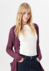 Blonde vrouw met golvend haar, gekleed in een witte geribbelde top, paarse cardigan en donkere denim jeans met haar handen in de zakken.