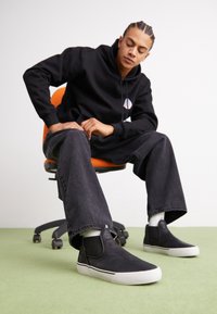 Chaussures noires à enfiler avec dessus en cuir texturé et semelle en caoutchouc blanche ; portées avec un jean noir taille haute et des chaussettes blanches visibles.