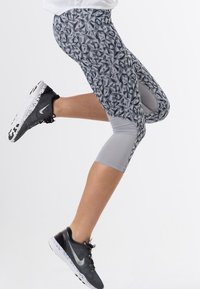 Šedé capri legíny s geometrickým designem a šedou jednobarevnou panely, doplněné černými sportovními botami s bílými akcenty.