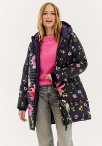 Černá květovaná péřová bunda s barevnými květinovými vzory, vybavená zipem, kapucí a žebrovanými manžetami, nošená přes růžový svetr.