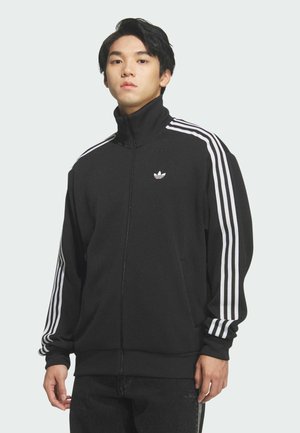 Młody mężczyzna w czarnej kurtce dresowej Adidas z białymi paskami na rękawach, stojący na jednolitym tle.