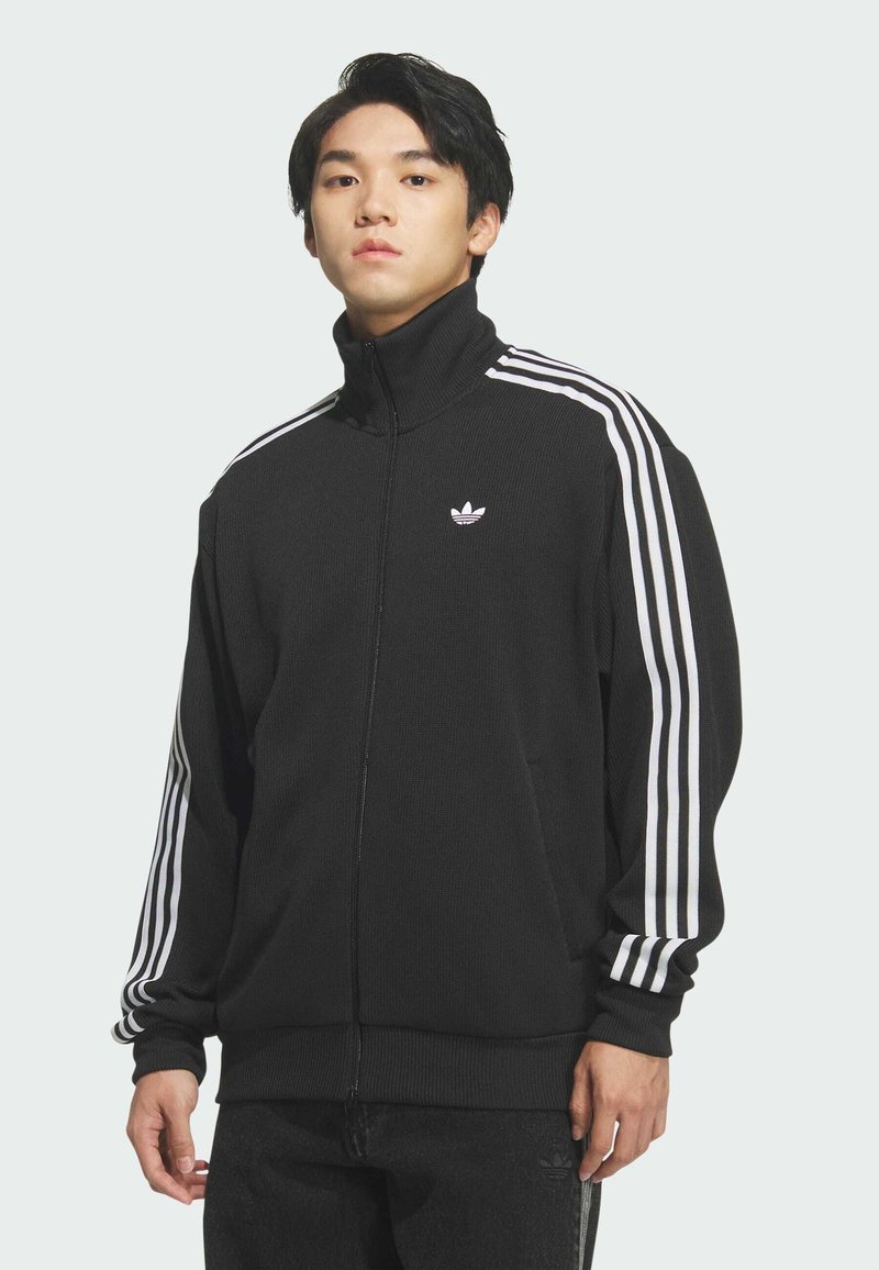 Jonge man draagt een zwart Adidas trainingsjack met witte strepen langs de mouwen, staand voor een effen achtergrond.