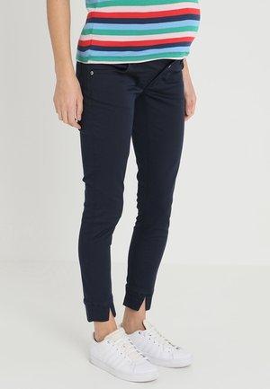 Personne portant un pantalon bleu marine foncé ajusté avec de petites fentes aux chevilles, des baskets blanches et une chemise rayée avec des bandes vertes, rouges, bleues et blanches.