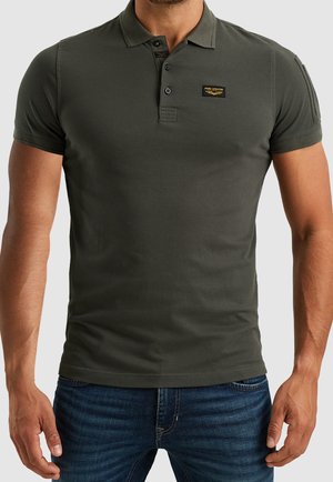 Man wearing fitted dark green short-sleeve polo shirt with three buttons and small logo patch, paired with blue jeans.