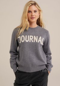 Szürke kötött pulóver "JOURNAL" felirattal krém színben, kerek nyakú, bordás mandzsettákkal és aljjal, puha textúrával, fekete nadrággal párosítva.