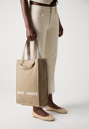 Beige canvas totebag med hvit tekst som sier "SAMSOE." Har solide håndtak og en strukturert form, kombinert med lyse bukser.