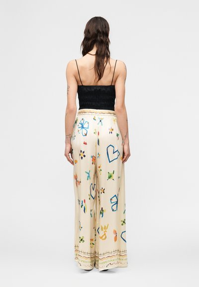 Γυναίκα με μακριά σκουρόχρωμα μαλλιά, που φοράει μαύρο αμάνικο τοπ και φαρδιά εκρού παντελόνια με πολύχρωμα αφηρημένα σχέδια, στέκεται με την πλάτη στραμμένη.