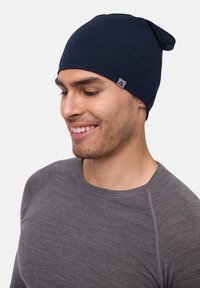 Marineblaue Beanie-Mütze mit spitzem Oberteil, aus geripptem Strickstoff, mit einem kleinen grauen Logotag an der Seite. Tragen mit einem grauen Langarmoberteil.