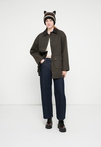 Πανωφόρι σε υπερμεγέθη γραμμή σε ελιά πράσινο χρώμα με δύο τσέπες μπροστά, φορεμένο πάνω από λευκή μπλούζα και σκούρο μπλε τζιν με φαρδιά παντελόνια. Μαύρα μποτάκια με κορδόνια ολοκληρώνουν το σύνολο.