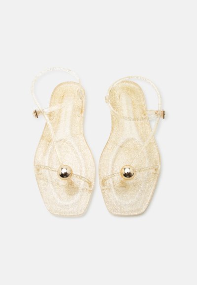 Sandale plate transparente sclipicioase, cu bretele subțiri, catarame la gleznă și un singur ornament rotund auriu pe fiecare bretea din față.