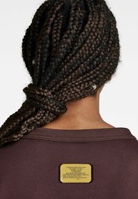 Ciemnobordowa bluza z okrągłym dekoltem i widoczną fakturą. Złota naszywka z logo przymocowana na tylnej części kołnierza. Warkoczyki ułożone z tyłu.