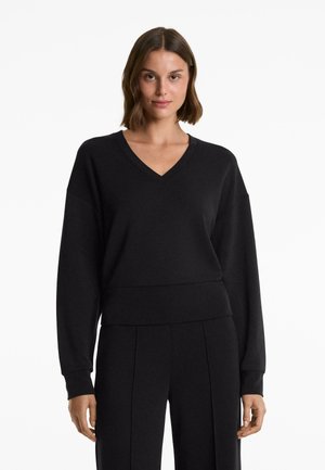 Woman with shoulder-length brown hair wearing a black V-neck long-sleeve sweater and black pants against a white background.