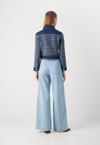 Veste en denim avec un motif texturé bleu et blanc, col et manches longues, assortie à un jean large bleu clair et des chaussures noires.