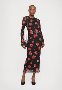 Lang svart kjole med rødt blomsterprint, tettsittende design, lange ermer og bølget kant. Sammen med en liten svart kjedehåndveske.