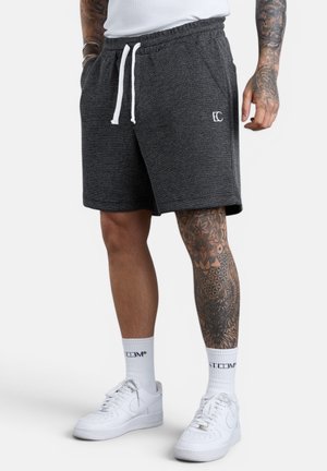 Mann trägt dunkelgraue, strukturierte Shorts mit weißen Kordeln, weißen Sportsocken, weißen Turnschuhen sowie tätowiertem Bein und Arm vor schlichtem Hintergrund.