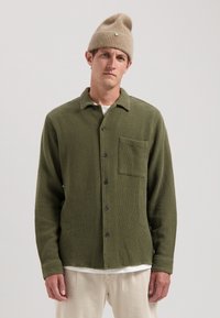 Cămașă butonată texturată verde, cu un buzunar pe piept, butoane negre și un guler casual, asortată cu o căciulă tricotată bej.