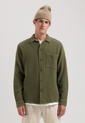 Green textured button-up shirt featuring a chest pocket, black buttons, and a casual collar, paired with a beige knit beanie.