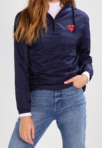 Person wearing navy blue half-zip windbreaker with red heart logo on chest and light blue jeans, hand resting in pocket.