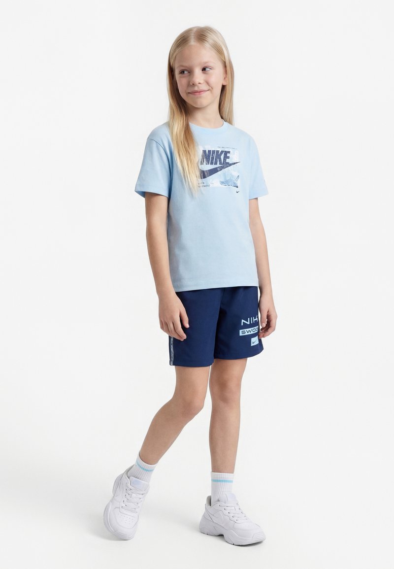 Noor tüdruk seisab, kandes helesinist Nike T-särki, tumesiniseid Nike lühikesi pükse, valgeid sokke ja valgeid tossusid ühevärvilise valge taustaga.