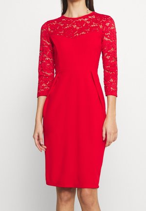 Rotes knielanges Kleid mit floraler Spitze auf der oberen Brust und dreiviertel Ärmel, getragen von einer Frau, die vor einem schlichten Hintergrund steht.