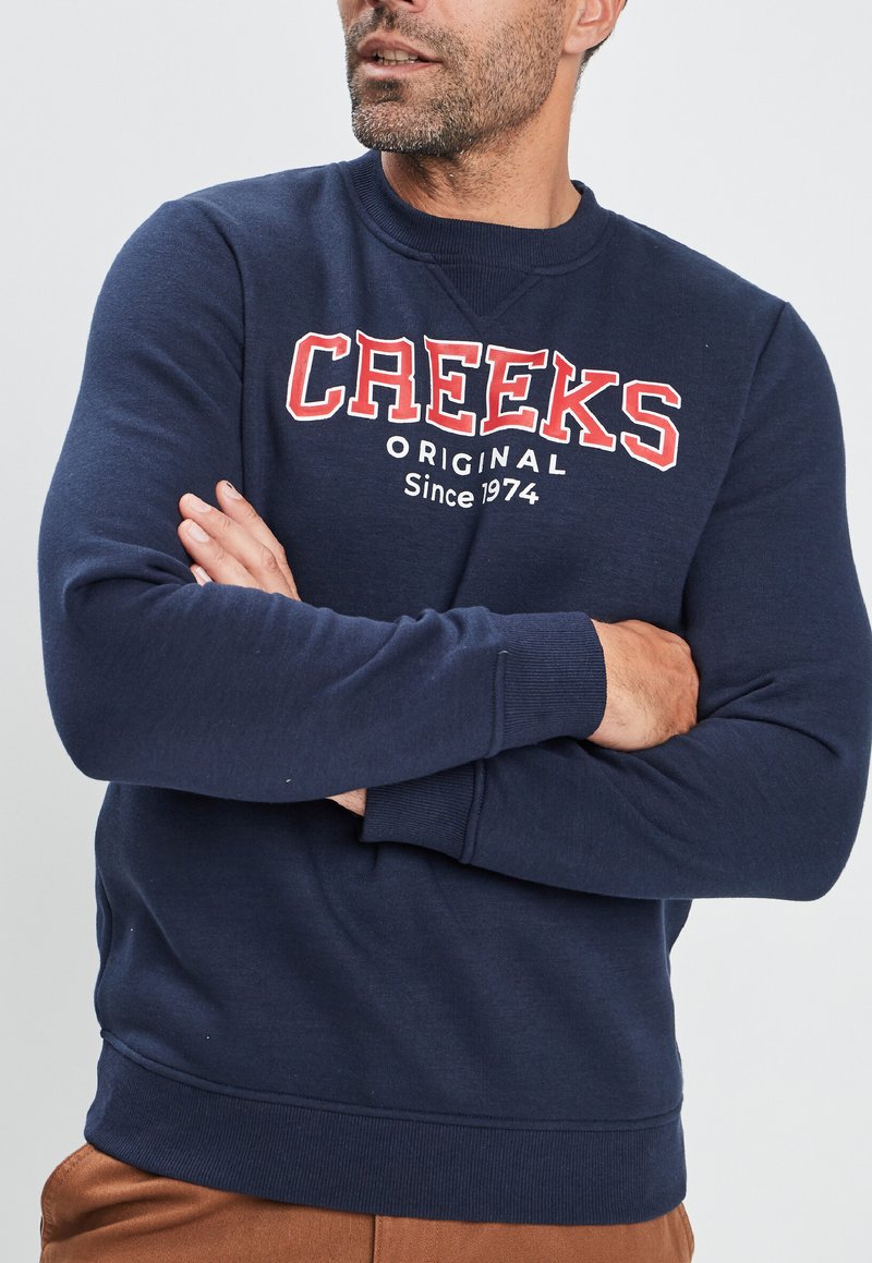 Sudadera azul marino de tejido suave, con un estampado en rojo y blanco que dice "CREEKS Original Desde 1974" en el pecho. Diseño clásico de cuello redondo.