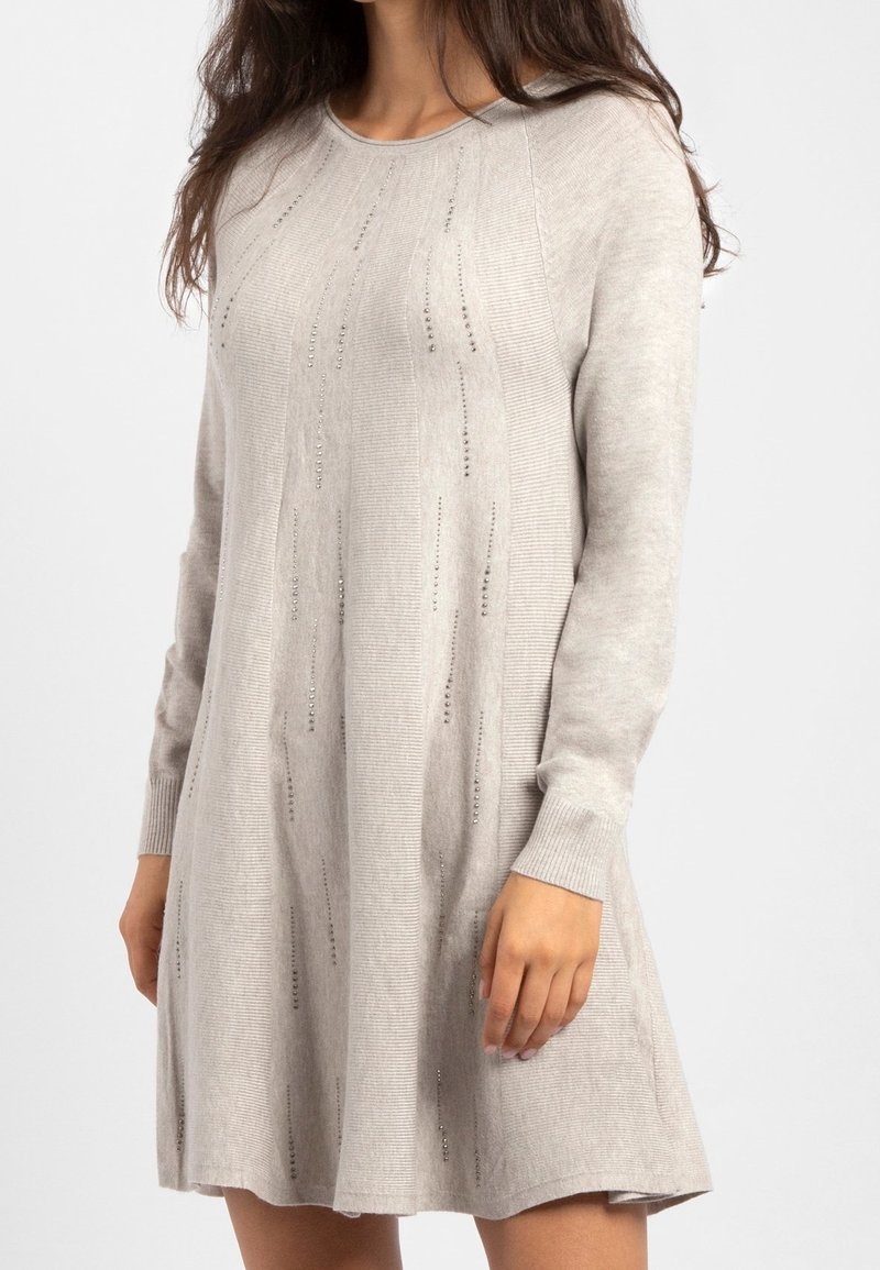 Femme portant une robe tricotée beige clair à manches longues avec des motifs décoratifs en perles verticales, se tenant avec un bras détendu à ses côtés.