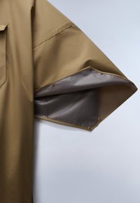 Cămașă maro supradimensionată cu o deschidere largă a mânecii, având o căptușeală interioară gri contrastantă și închideri cu capse pe margine.