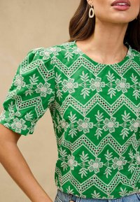 Blusa verde con maniche a sbuffo corte, caratterizzata da ricami floreali bianchi e motivi intricati. Collo rotondo e tessuto leggero.