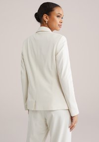 Moteris su tamsiais surištais plaukais, vilkinti kremine švarkelio ir derančių kelnių komplektu, matoma iš nugaros priešais vienodą šviesią foną.