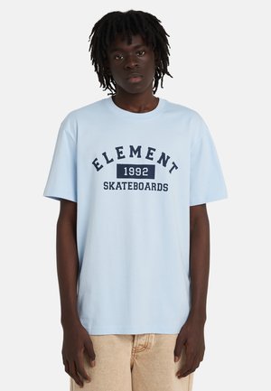 Młoda osoba z dredami, ubrana w jasnoniebieską koszulkę Element 1992 Skateboards i beżowe spodnie, stojąca na białym tle.