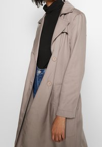 Trench coat color taupe chiaro con design su misura, bottoni doppi, e una texture morbida, indossato sopra un dolcevita nero.
