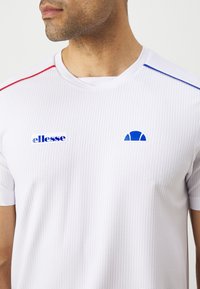 Valge lühikeste varrukatega spordipaita, millel on tekstureeritud muster, sinised ja punased aktsendid ning logod "ellesse" ja poolringikujuline disain.