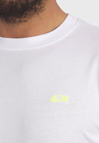 T-shirt bianco in cotone con collo rotondo, dotato di un piccolo logo giallo neon sul petto. Il tessuto appare liscio e leggero.