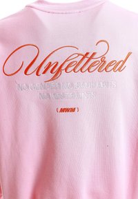 Sudadera rosa con texto bordado. Al frente dice "Unfettered" en escritura naranja, con texto blanco adicional debajo. Textura suave.