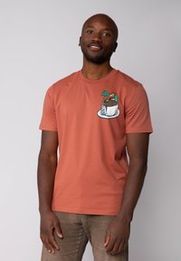 T-shirt en coton couleur rouille avec un design brodé représentant une tasse avec un dessert et des palmiers sur le côté gauche de la poitrine.