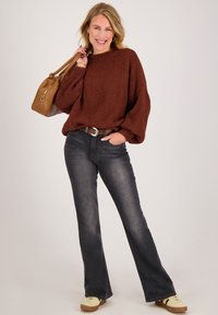 Vriendelijke vrouw met een bruine grove trui, zwarte uitlopende jeans, gele sneakers, een bruine handtas over de schouder, poserend tegen een effen achtergrond.