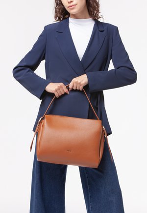 Borsa a mano in pelle marrone con una forma strutturata, dotata di chiusura a zip e una singola maniglia, tenuta da una persona in giacca blu navy.